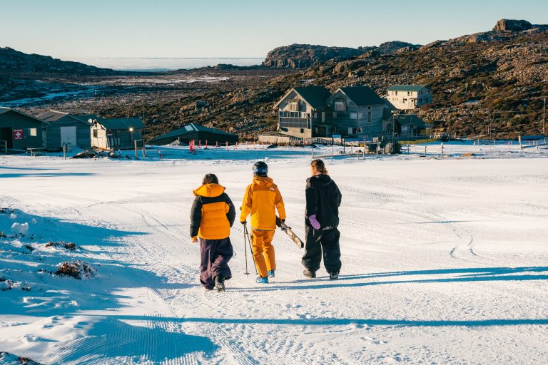 3 people dressed in winter clothing walking through Ben Lomond Alpine resort with ski and snowboard gear.